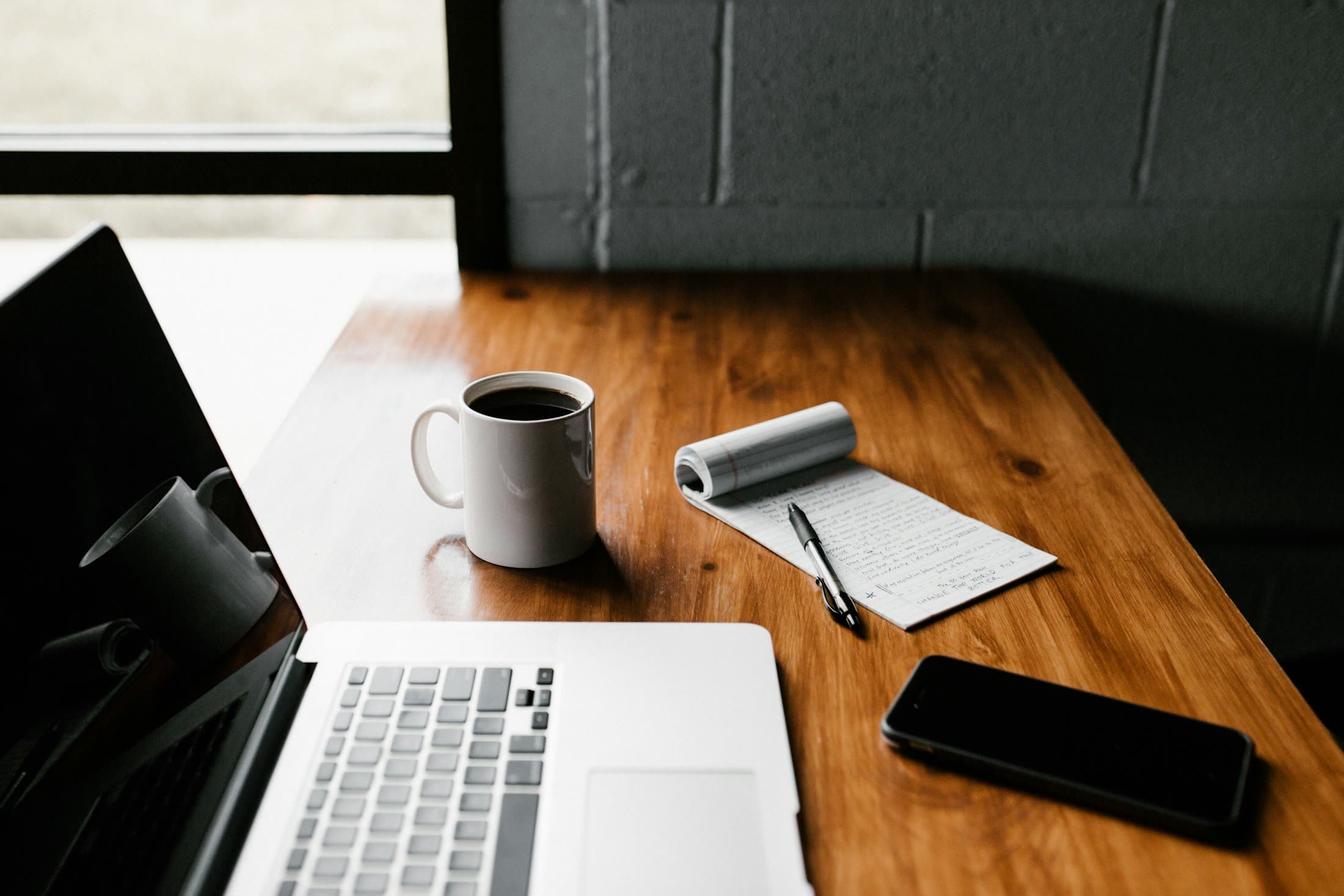 A laptop, coffee and notebook on a warm wooden desk